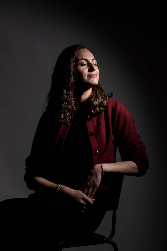 Chaya Koritz sitting peacefully on a chair, softly lit with gentle light on her face, wearing a red jacket.