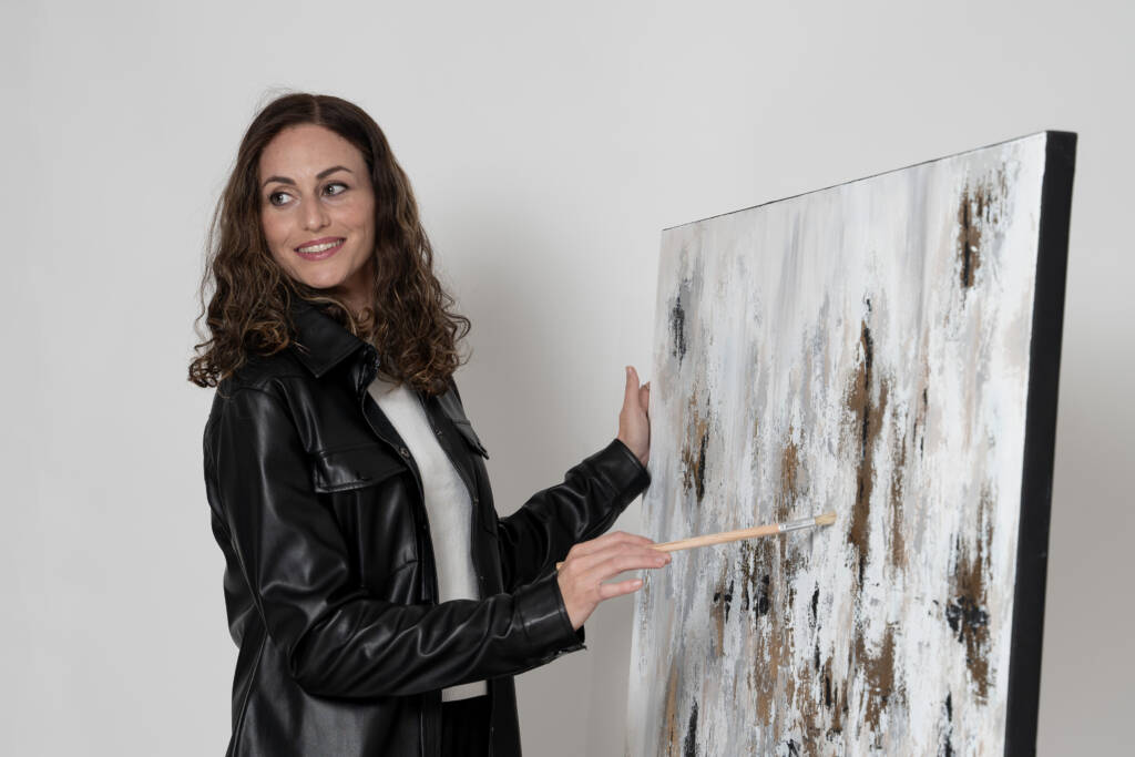 Chaya Koritz smiling warmly while painting next to her artwork, immersed in the creative process.