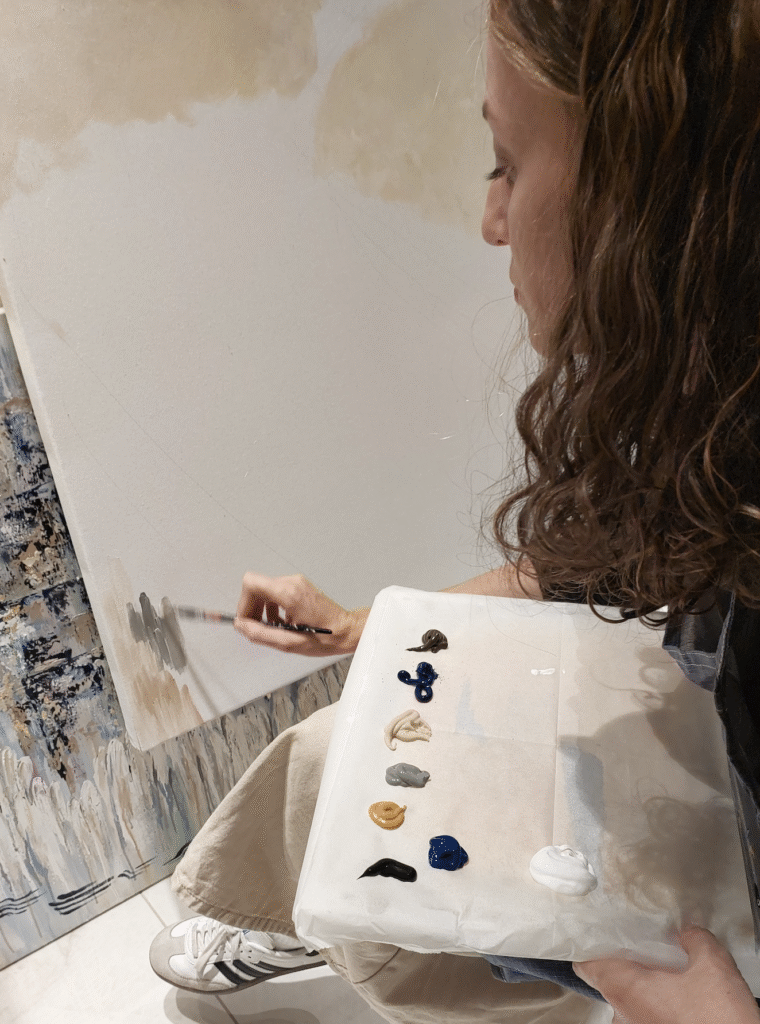 Chaya is holding a palete in her left hand and a paintbrush in her right hand. She is leaning over her canvas, starting a new work of art.