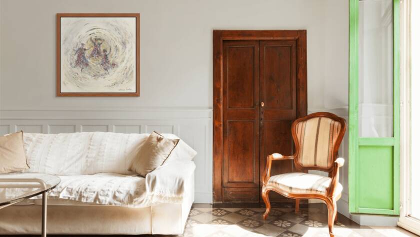 Framed artwork of three Hasidic men dancing in a golden circle, hanging above a modern white sofa beside a vintage wooden door and sunlit window.