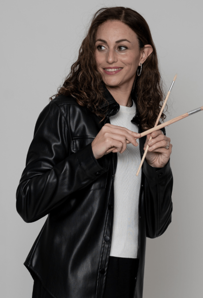 Chaya Koritz – Artist Holding Paintbrush with Warm Smile Chaya Koritz holding a paintbrush with a warm, friendly smile, radiating creativity and passion for art.